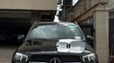 Front view of a black Mercedes SUV parked in a driveway, with buildings and a barbed-wire fence nearby, water tanks on the roof area in the background.