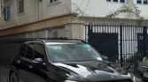 Black Mercedes SUV parked in front of a gated building with barbed wire and a white two-story house with blue-framed windows behind it.