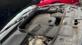 Open hood shows a dusty engine bay of a silver/gray SUV with other parked cars in the background, including a red car closest behind the engine bay.