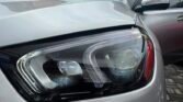 Close-up of a silver car's front-left headlight with the hood slightly raised and a driver-side door open in a residential driveway backdrop.
